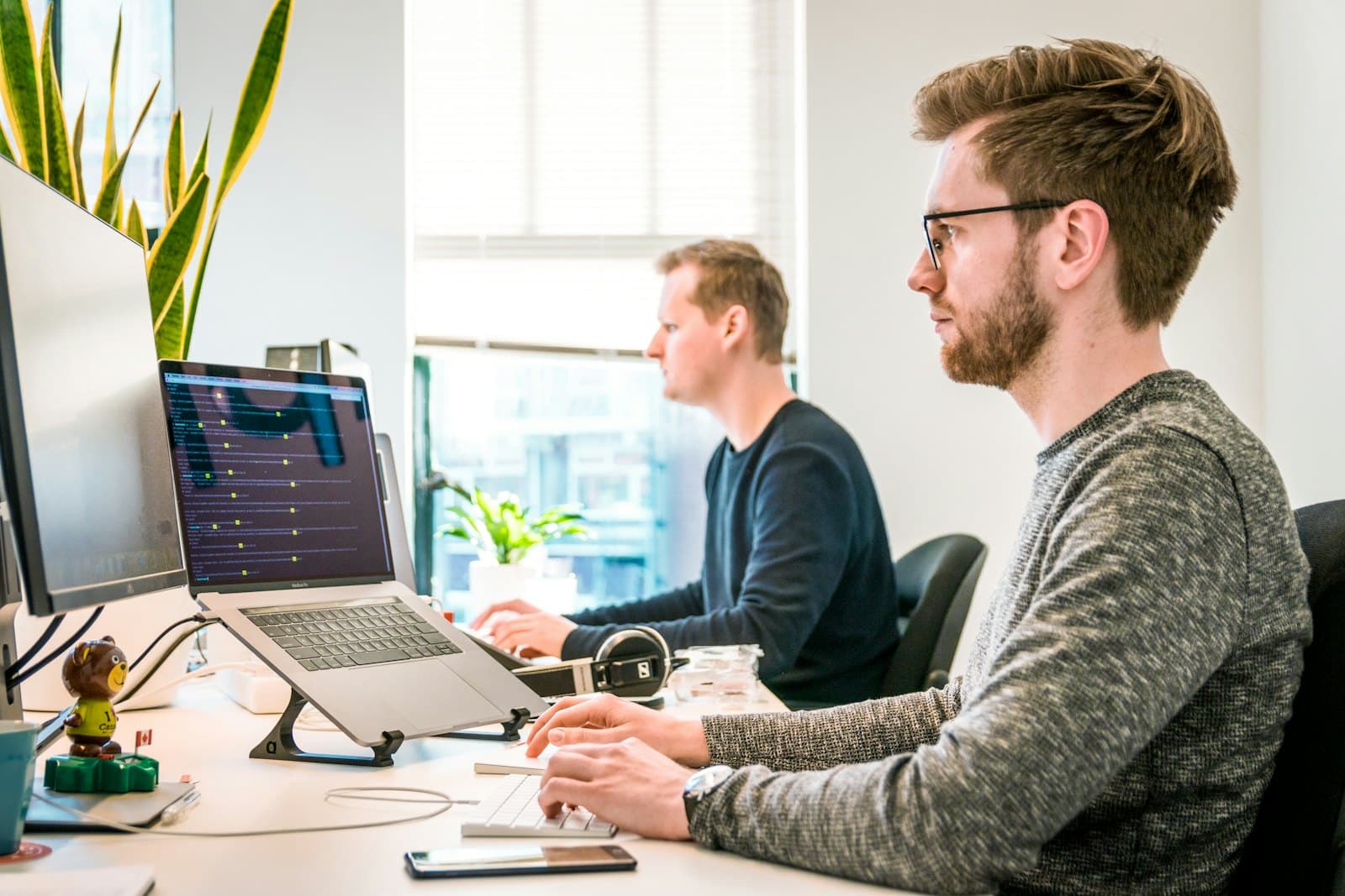 Two engineers at a shared desk, one reviewing code on a laptop screen